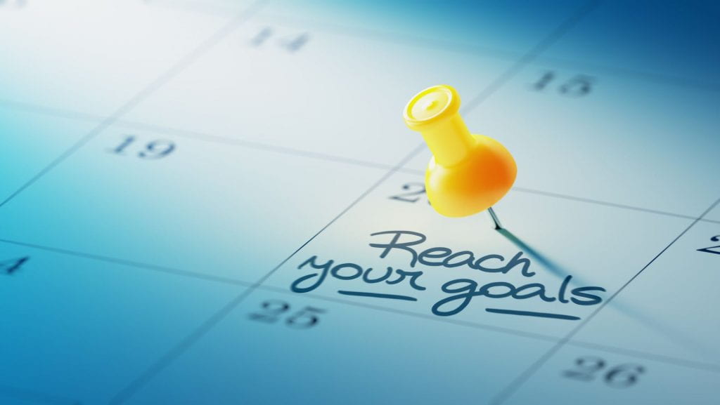 A calendar with a push pin placed on a date with the reminder ''reach your goals'', emphasizing the importance of following a calendar to predict stressors on college students.